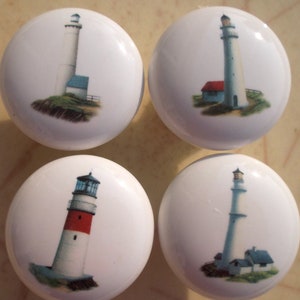May include: Four white ceramic cabinet knobs with lighthouse designs. Each knob features a different lighthouse illustration, with varying colors and architectural details. The knobs are round and have a glossy finish, suitable for furniture or decor.