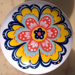 May include: White ceramic cabinet knob with a colorful floral design. The flower features a yellow, red, blue, and teal pattern. The knob is round and has a glossy finish, suitable for drawers or cabinets.