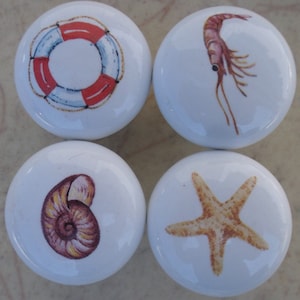 May include: Set of four ceramic knobs with nautical themes. Each knob features a different design: a red and white life preserver, a brown shrimp, a brown seashell, and a tan starfish.