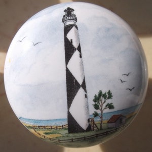 May include: White ceramic knob featuring a lighthouse with black and white diamond pattern. The knob has a coastal scene with a small house, tree, and birds against a blue sky and ocean background. The knob is round.