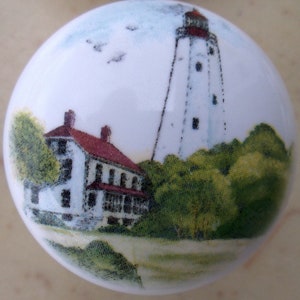 May include: A white ceramic knob with a painted image of a lighthouse, a house, and trees. The lighthouse is white with a red roof and is surrounded by green trees. The house is white with a red roof and is located in front of the lighthouse. The knob is a great addition to any nautical themed home.