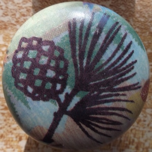 May include: A round, fabric-covered button with a brown pine branch and pine cone design on a light blue, green, and brown background.