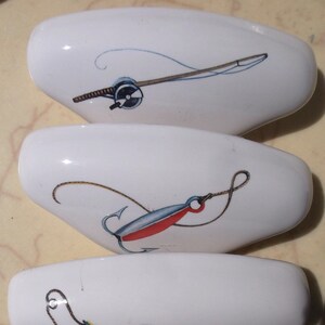 May include: Three white ceramic knobs with fishing-themed illustrations. The top knob features a fishing rod and reel, the middle knob shows a lure with a hook, and the bottom knob has a colorful lure with a hook.
