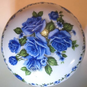 May include: A white, round decorative object featuring a cluster of blue roses with green leaves. The roses are the focal point, with smaller blue flowers and a delicate blue floral border. The object has a glossy finish.