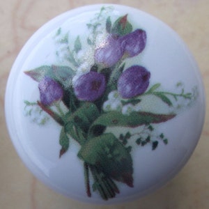 May include: A white ceramic knob with a floral design. The design features a bouquet of purple flowers with green leaves and white flowers.
