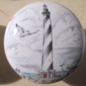 May include: A white ceramic knob with a painted image of a black and white striped lighthouse with a seagull flying in the sky. The lighthouse is set against a blue sky with white clouds.