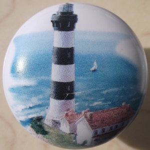 May include: A white ceramic knob with a black and white lighthouse image. The lighthouse is on a cliff with a blue ocean and a sailboat in the background.