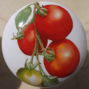 May include: A white ceramic knob with a painted image of red tomatoes on a vine. The tomatoes are arranged in a cluster, with one larger tomato at the top and two smaller tomatoes below. The vine is green and has leaves.
