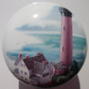 May include: A ceramic knob with a painted image of a pink lighthouse with a white top and a blue sky with white clouds. There are also some small houses in the foreground.