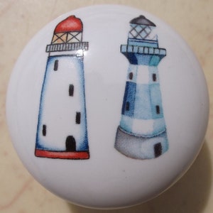 May include: White ceramic cabinet knob featuring two lighthouse designs. One lighthouse has a red roof, the other a black roof. Both lighthouses are blue and white with black windows. The knob is round.