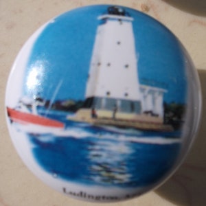 May include: A white lighthouse with a red boat in front of it. The lighthouse is on a small island with a blue sky and water in the background. The text "Ludington, MI" is at the bottom of the image.