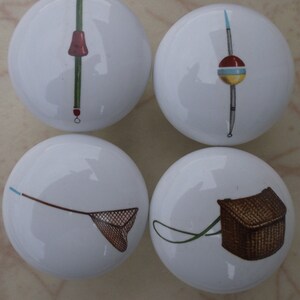 May include: Four white ceramic knobs with fishing-themed designs. The knobs feature a fishing rod with a bobber, a fishing net, a fishing basket, and a fishing rod with a lure.