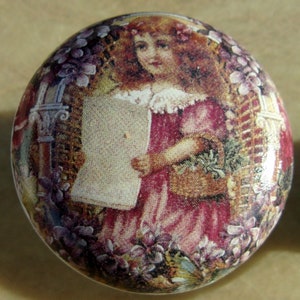 May include: A round, ceramic knob with a vintage image of a girl in a pink dress holding a basket of flowers. The background is a floral pattern with a trellis and a stone arch.