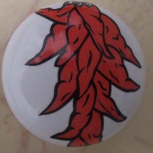May include: A white ceramic cabinet knob featuring a cluster of red chili peppers with black outlines. The peppers are arranged in a downward-facing pattern. The knob is round and has a glossy finish.