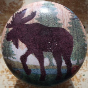May include: A round cabinet knob featuring a dark purple moose silhouette against a backdrop of a forest scene. The design includes green trees, a blue river, and a light-colored sky, creating a rustic aesthetic.