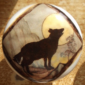 May include: A white ceramic knob with a brown and beige design of a wolf howling at the moon. The wolf is a silhouette against a yellow moon.
