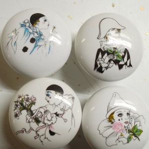 May include: Four white ceramic cabinet knobs with illustrations of Pierrot figures in various poses. Each knob features a different design, including a figure with a flower and another blowing a dandelion.