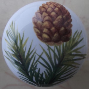May include: A white ceramic knob with a painted image of a brown pine cone and green pine needles.