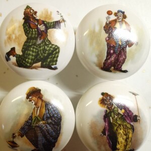May include: Four round white ceramic cabinet knobs, each featuring a detailed illustration of a clown in a different pose and colorful outfit. The clowns are depicted with painted faces, wearing patterned clothing in shades of green, purple, blue, and yellow, and performing various activities.