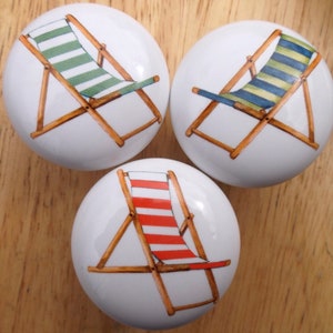 May include: Three white ceramic cabinet knobs with beach chair designs. One knob features a green and white striped chair, another has blue and yellow stripes, and the third has red and white stripes. The knobs have a wooden frame.