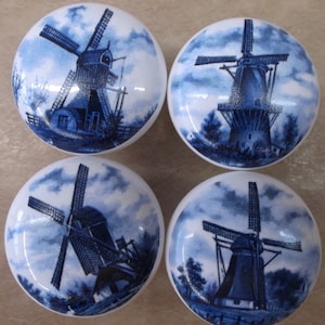 May include: Set of four white ceramic cabinet knobs with blue windmill illustrations. Each knob features a different windmill scene against a cloudy sky. The knobs are round and have a glossy finish, suitable for furniture.