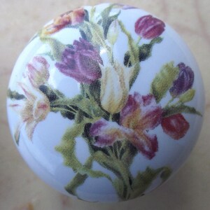 May include: A white ceramic cabinet knob featuring a floral design. The flowers are a mix of purple, yellow, and red, with green stems and leaves. The knob is round and has a glossy finish.