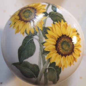 May include: A white ceramic knob with a sunflower design. The knob features two sunflowers with yellow petals and brown centers, along with green leaves.