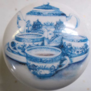 May include: White ceramic knob featuring a blue and white illustration of a teapot and teacups. The design depicts a traditional tea set with detailed landscape scenes. The knob is round and has a glossy finish, suitable for furniture or cabinet hardware.