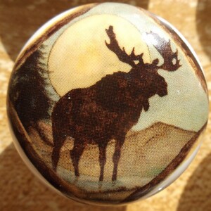 May include: A ceramic knob with a brown silhouette of a moose standing in front of a yellow moon on a beige background.