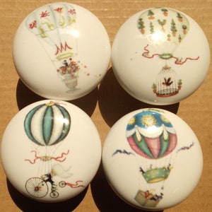 May include: Four white ceramic cabinet knobs with colorful hot air balloon designs. Each knob features a different balloon illustration, including floral arrangements, a birdcage, and a bicycle. These decorative knobs add a whimsical touch to furniture.