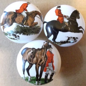 May include: Three oval ceramic knobs with a white background and a hunting scene with a rider on a horse. The first knob features a brown horse jumping over a fence. The second knob features a black horse with a rider. The third knob features a brown horse standing next to a rider.