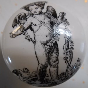 May include: A black and white ceramic tile with a detailed image of a winged cherub holding a lyre. The cherub is surrounded by floral designs.