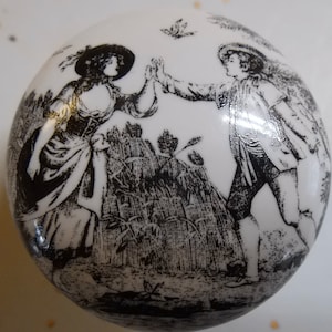 May include: Black and white ceramic knob with a design of two people in a field of wheat. The people are dressed in 18th-century clothing and are holding hands.