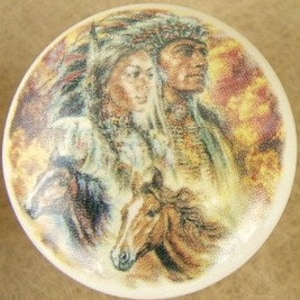 May include: A round, white ceramic knob featuring a detailed illustration of a Native American couple and two horses. The artwork uses warm tones of brown, gold, and beige, with a soft, painterly style. The knob is likely for furniture or decor.