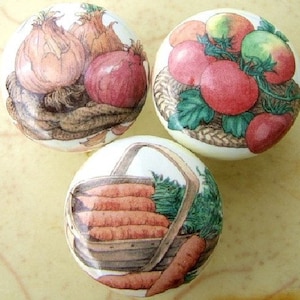 May include: Set of three ceramic knobs featuring a vintage style illustration of vegetables. One knob shows onions, another shows tomatoes, and the last shows carrots in a basket.