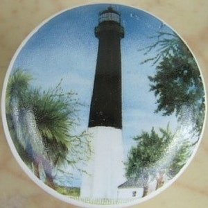 May include: A white and black lighthouse with a white base and a black tower, surrounded by green trees against a blue sky.