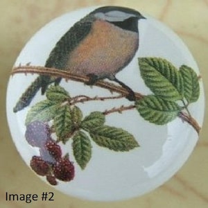 May include: A white ceramic round ornament with a black-capped chickadee perched on a branch with green leaves and purple and red berries.