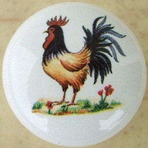 May include: White ceramic cabinet knob featuring a colorful rooster illustration. The rooster has black, brown, and yellow feathers, a red comb and wattle, and a black tail. Small red flowers and green grass are at the base.