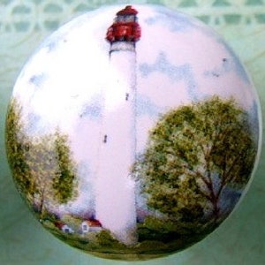 May include: A round, white ornament featuring a painted lighthouse scene. The lighthouse is white with a red top, set against a backdrop of blue sky and green trees. A small house is visible at the base of the lighthouse.