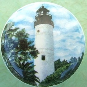 May include: A white lighthouse with a black roof and a black railing stands tall against a blue sky with white clouds. Lush green trees surround the base of the lighthouse.