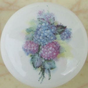 May include: A white ceramic knob with a floral design. The design features a bouquet of purple and blue flowers with green leaves.