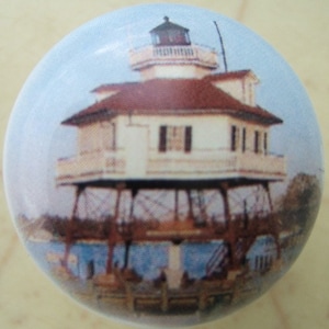 May include: A round decorative knob featuring a detailed illustration of a lighthouse. The lighthouse has a white exterior, a red roof, and a black lantern room. The background is a light blue sky and water scene.