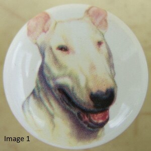 May include: A white ceramic knob with a painted image of a white bull terrier dog with pink ears and a black nose. The dog is looking to the right and has its mouth open, showing its tongue.