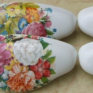 May include: Four white oval-shaped ceramic drawer pulls. Each pull is decorated with a vibrant floral arrangement featuring pink, yellow, blue, and orange flowers. The pulls are arranged in a square formation.
