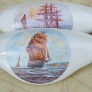 May include: Four white, oval-shaped decorative stones. Each stone features a circular image of a tall sailing ship on the ocean, with brown sails and a blue and white sky. The stones have a glossy finish.