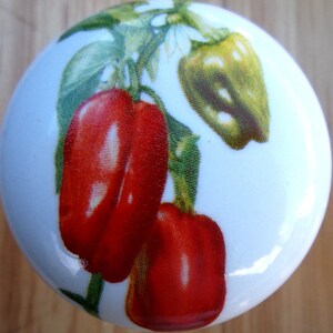May include: A white ceramic knob with a red and green pepper design. The peppers are arranged vertically on a vine with green leaves.