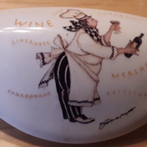 May include: White ceramic wine bottle stopper with a cartoon chef holding a glass of wine and a bottle of wine. The stopper is decorated with the words "Wine", "Zinfandel", "Chardonnay", "Merlot", and "Riesling".