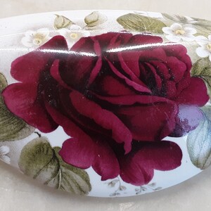 May include: Oval-shaped, white ceramic trinket box featuring a large, deep red rose as the central design element. The rose is surrounded by green leaves and small white flowers, creating a floral pattern. The box has a glossy finish.