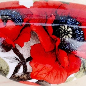 May include: Oval-shaped decorative ceramic knob featuring a vibrant red poppy floral design. The poppy has black and blue accents, with green leaves on a white background. This decorative knob is ideal for cabinets and drawers.