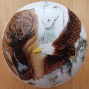 May include: White ceramic cabinet knob featuring a brown bear, a white horse, a bald eagle, and a gray wolf. The knob has a glossy finish and is decorated with detailed illustrations of the animals.